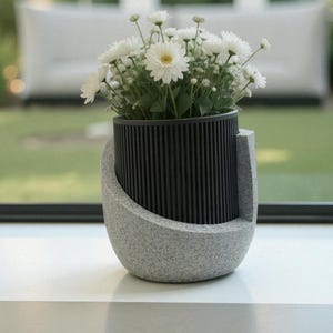May include: A contemporary flower pot in grey and black, filled with white daisies. The pot features a textured, curved base and a black, vertically-lined cylindrical section. The background shows a window and a white sofa.
