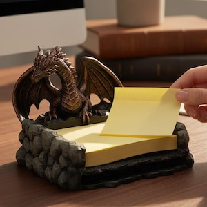 May include: A bronze-colored dragon-shaped memo pad holder with a stack of yellow sticky notes. The dragon sits atop a stone-like base. A person is seen taking a sticky note from the holder. A computer, mug, and plant are in the background.