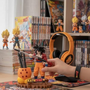 Op de afbeelding: Een bureauscène met Dragon Ball Z-merchandise. Inclusief beeldjes, een ingelijste poster en een laptop. Een bureau-organizer met de naam "KATHERINE" is zichtbaar. Een oranje bol met sterren en een oranje koptelefoon zijn ook aanwezig.
