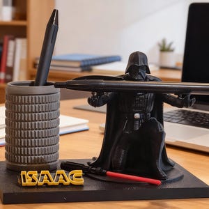 以下が含まれることがあります： 黒いダース・ベイダーのペン立て。ペンとライトセーバーが付属。ペン立ては円筒形で、黒いペンが入っています。ベースには黄色い文字で「ISAAC」と書かれています。背景にはノートパソコンがあります。