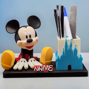 May include: A Mickey Mouse desk tidy. The black, red, white, and yellow figure sits beside a white and blue castle-shaped pen holder. The base is black, and a red and white sign reads "NAME". Pens and a nail file are inside.