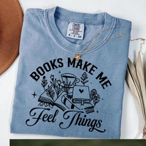 May include: A light blue t-shirt with the text "BOOKS MAKE ME Feel Things" in black script. The graphic includes illustrations of books, flowers, and a coffee cup. A gold necklace rests on the shirt.
