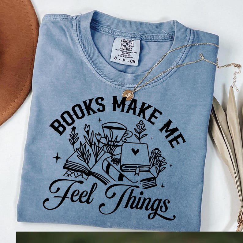 May include: A light blue t-shirt with the text "BOOKS MAKE ME Feel Things" in black script. The graphic includes illustrations of books, flowers, and a coffee cup. A gold necklace rests on the shirt.