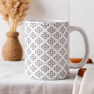 May include: White ceramic mug with a repeating black floral pattern. The mug has a curved handle and is set on a white tablecloth. A small vase with dried wheat and a decorative vase are in the background.