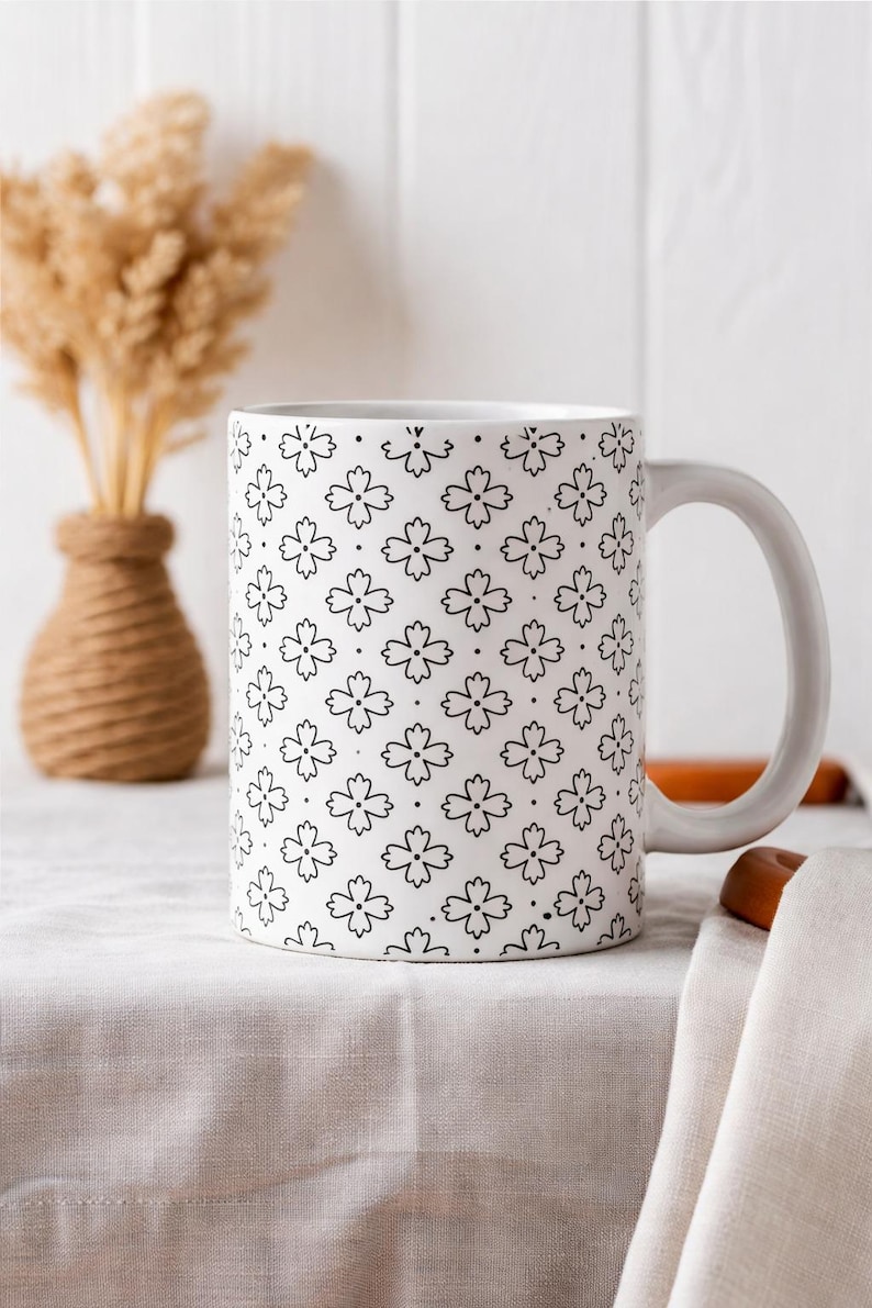 May include: White ceramic mug with a repeating black floral pattern. The mug has a curved handle and is set on a white tablecloth. A small vase with dried wheat and a decorative vase are in the background.