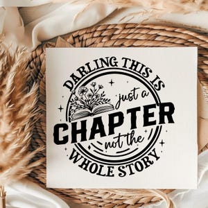 May include: White square print with the text "DARLING THIS IS just a CHAPTER not the WHOLE STORY" in a black circular design. The design includes an open book with floral accents. The print is on a white surface.