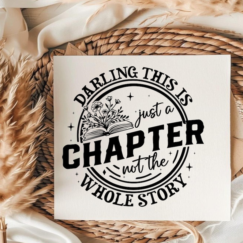 May include: White square print with the text "DARLING THIS IS just a CHAPTER not the WHOLE STORY" in a black circular design. The design includes an open book with floral accents. The print is on a white surface.