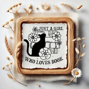 May include: A framed print with the text "JUST A GIRL WHO LOVES BOOK" and a black cat silhouette atop a stack of books, surrounded by floral accents. The frame is a light brown color, and the background is a neutral beige.