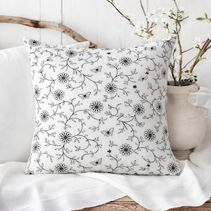 May include: A white square pillow with a black floral pattern featuring daisies, butterflies, and vines. The pillow is placed on a white surface, with a white vase of flowers in the background.
