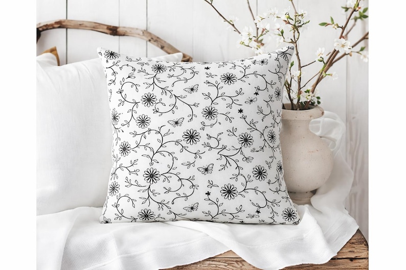 May include: A white square pillow with a black floral pattern featuring daisies, butterflies, and vines. The pillow is placed on a white surface, with a white vase of flowers in the background.
