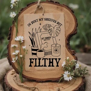 May include: A rustic wooden sign with the text "IN DUST MY SHELVES BUT MY BOOKS ARE STILL FILTHY" in black. The sign features illustrations of books, plants, a candle, and a coffee cup. The sign is displayed on a wooden slice.
