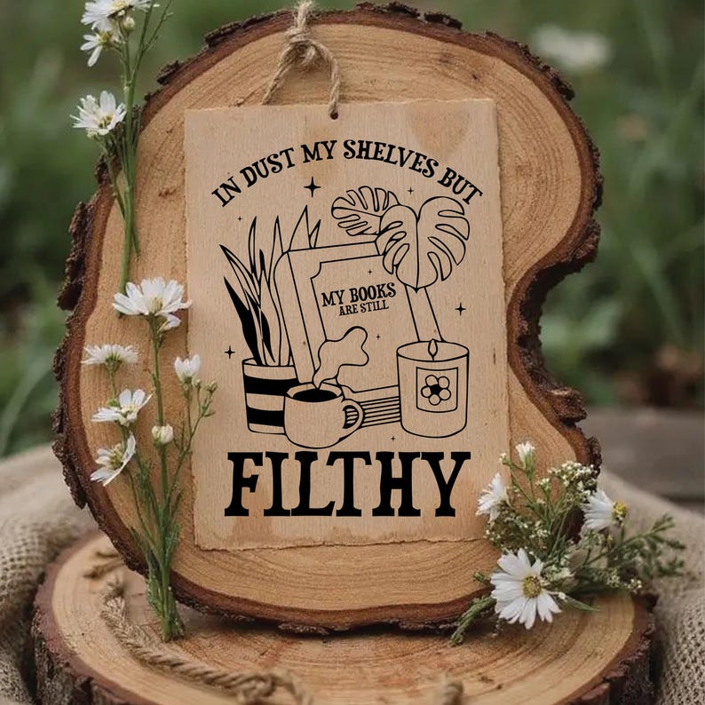May include: A rustic wooden sign with the text "IN DUST MY SHELVES BUT MY BOOKS ARE STILL FILTHY" in black. The sign features illustrations of books, plants, a candle, and a coffee cup. The sign is displayed on a wooden slice.