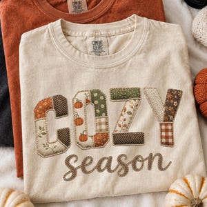 Puede incluir: Tres camisetas de cuello redondo en negro, naranja óxido y crema. La camiseta crema presenta el texto COZY season en una fuente estilo patchwork con patrones otoñales que incluyen calabazas, hojas y cuadros. Cada camiseta tiene una etiqueta Comfort Colors.