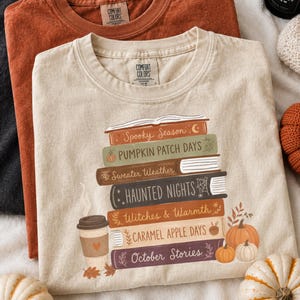 Puede incluir: Tres camisetas de cuello redondo en negro, óxido y crema. La camiseta crema presenta un gráfico de una pila de libros con los títulos Spooky Season, Pumpkin Patch Days, Sweater Weather, Haunted Nights, Witches & Warmth, Caramel Apple Days y October Stories.