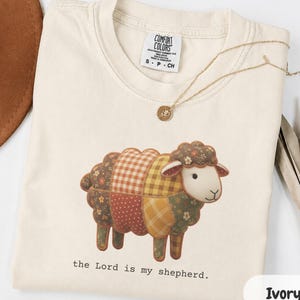 May include: An ivory t-shirt featuring a graphic of a sheep with a patchwork body made of various brown, yellow, and red floral and plaid patterns. Below the sheep, the text reads, "the Lord is my shepherd." A gold necklace rests on the collar.