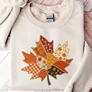 May include: A cream-colored crewneck sweatshirt featuring a large, central maple leaf graphic composed of various autumn-themed fabric patterns, including plaid, polka dots, and small pumpkin motifs in shades of orange, brown, and white.