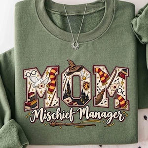 Peut inclure: Un sweat-shirt à col rond vert olive avec le texte MOM en grandes lettres majuscules remplies d'illustrations sur le thème des sorciers, dont une chouette blanche, des lunettes, une écharpe, un chapeau et un château. Le texte Mischief Manager est écrit en dessous.