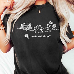 May include: Black t-shirt with a white graphic of books, a paw print, and a coffee cup. The text "My needs are simple" is below the graphic. The shirt has a crew neck and short sleeves.