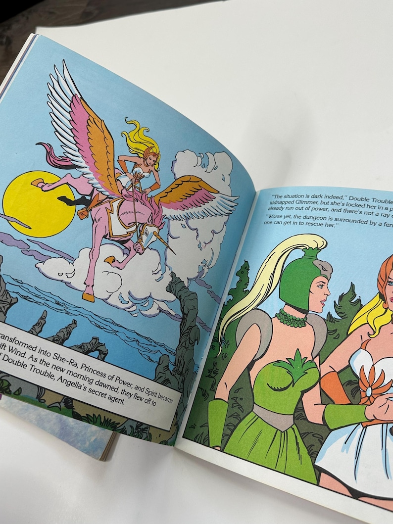 May include: Open book illustration featuring a blonde woman riding a pink unicorn with large pink and white wings. The background is blue with a yellow sun and white clouds. Text on the page reads "transformed into She-Ra, Princess of Power..."