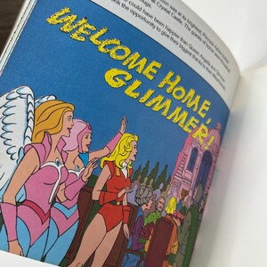 May include: An illustration from a children's book features the text "WELCOME HOME, GLIMMER!" in yellow letters against a blue sky. Several cartoon characters are depicted, including women in pink and red outfits, and a castle is visible in the background.