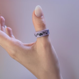 Op de afbeelding: Een lichtpaarse ring met een zwart bloemmotief, gedragen op een duim. Het ontwerp omvat rozen en bladeren. De ring is gemaakt van een flexibel materiaal.