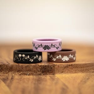 K&ouml;nnte beinhalten: Drei Silikonringe &uuml;bereinander gestapelt, jeweils mit einem floralen Design. Der oberste Ring ist lavendelfarben mit einem schwarzen Blumenmuster, der mittlere Ring ist schwarz mit einem silbernen Blumenmuster und der untere Ring ist braun mit einem wei&szlig;en Blumenmuster.