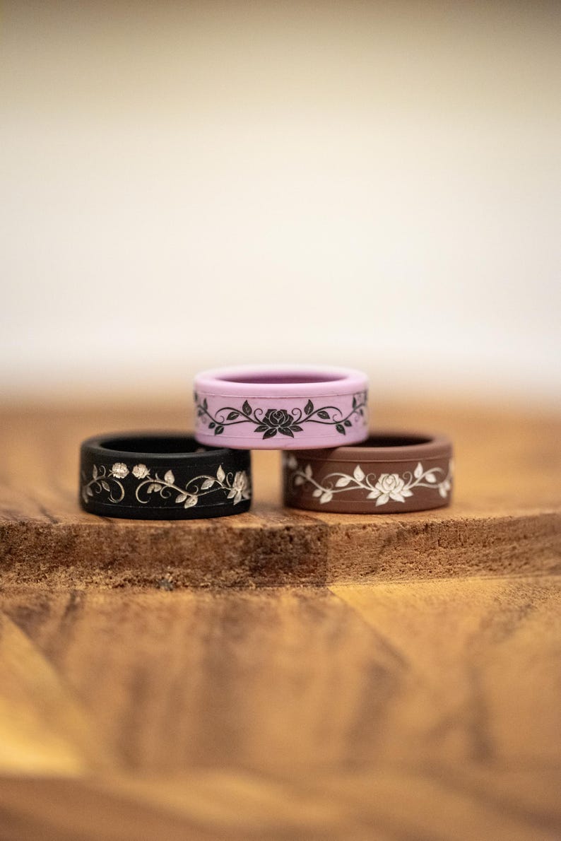 K&ouml;nnte beinhalten: Drei Silikonringe &uuml;bereinander gestapelt, jeweils mit einem floralen Design. Der oberste Ring ist lavendelfarben mit einem schwarzen Blumenmuster, der mittlere Ring ist schwarz mit einem silbernen Blumenmuster und der untere Ring ist braun mit einem wei&szlig;en Blumenmuster.