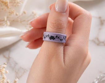 Könnte beinhalten: Ein lila Ring mit einem schwarzen Blumenmuster wird an einem Finger getragen. Der Ring hat eine glatte Oberfläche und ein Band mit einem sich wiederholenden Muster aus Rosen und Blättern. Der Hintergrund ist verschwommen, mit Andeutungen von weißem Stoff und dekorativen Elementen.
