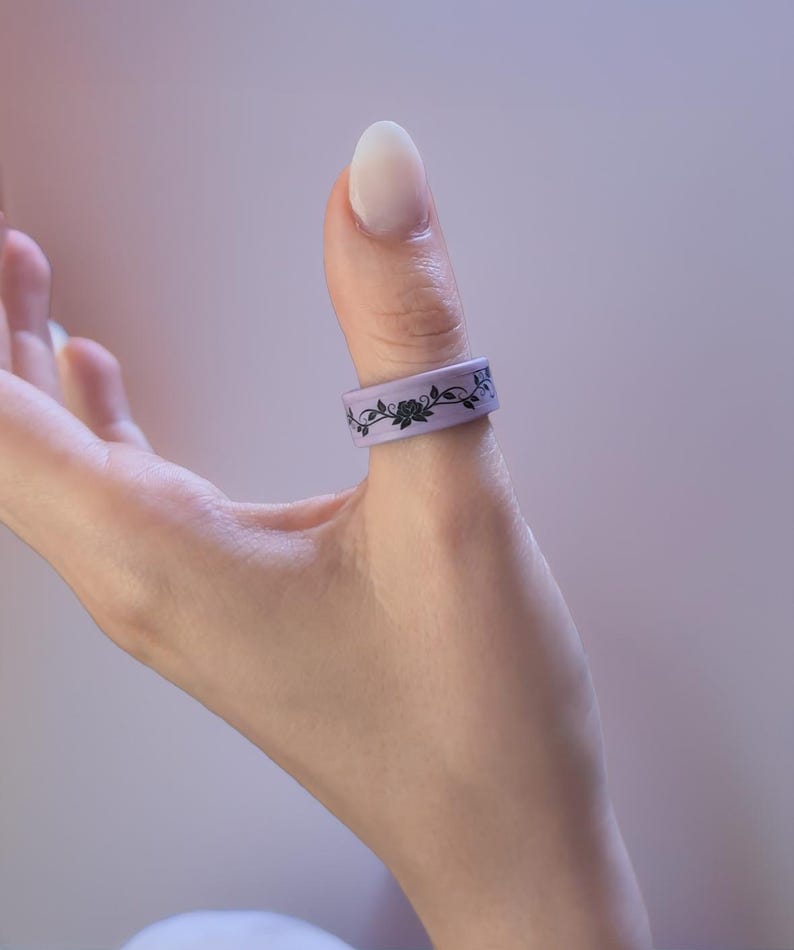 K&ouml;nnte beinhalten: Ein hellvioletter Ring mit einem schwarzen Blumenmuster, der an einem Daumen getragen wird. Das Design umfasst Rosen und Bl&auml;tter. Der Ring besteht aus einem flexiblen Material.