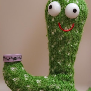 K&ouml;nnte beinhalten: Ein skurriles gr&uuml;nes Kaktus-Pl&uuml;schtier mit gro&szlig;en wei&szlig;en Augen und einem rot gestickten L&auml;cheln. Es tr&auml;gt einen kleinen schwarzen Hut mit floralen Details und h&auml;lt einen kleinen Beh&auml;lter mit einem floralen Design.