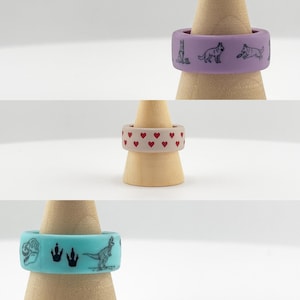 Op de afbeelding: Drie siliconen ringen op houten standaards. Eén is lavendelkleurig met hondenillustraties, een andere is beige met rode harten en de derde is turquoise met dinosaurusafbeeldingen.