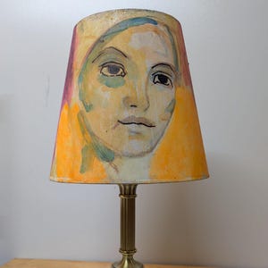 Może przedstawiać: Mosiężna lampa stołowa z malowanym abażurem. Abażur przedstawia akwarelowy portret kobiety w żółtych, pomarańczowych i fioletowych odcieniach. Lampa ma okrągłą podstawę i żłobkowaną kolumnę.