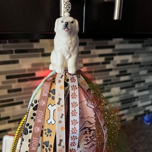 Peut inclure: Figurine de chien en céramique blanche souriante, surmontant un pompon coloré. Le pompon présente des rubans avec des empreintes de pattes, des os de chien et le mot "WOOF". Un charm argenté et des perles sont en haut.