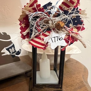 May include: A decorative lantern with a battery-operated candle. The lantern is adorned with a patriotic bow featuring red, white, and blue fabric. A star-shaped tag with the number "1776" is attached. The lantern sits on a wooden surface.
