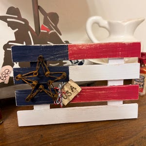 May include: A wooden American flag decoration with red, white, and blue painted stripes. A star made of twisted wire is attached to the blue section. A small tag with "USA" is tied to the flag.