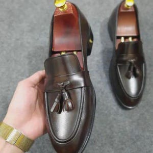 May include: A pair of dark brown leather loafers featuring a tassel detail on the vamp and a wooden shoe tree inserted into the shoe held in the hand. The loafers have a smooth finish, a rounded toe, and a low heel, showcasing a classic slip-on design.