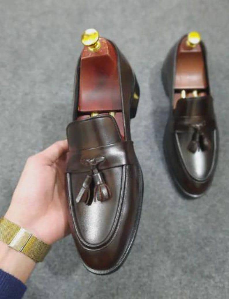 May include: A pair of dark brown leather loafers featuring a tassel detail on the vamp and a wooden shoe tree inserted into the shoe held in the hand. The loafers have a smooth finish, a rounded toe, and a low heel, showcasing a classic slip-on design.