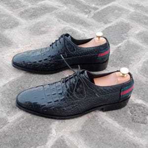 K&ouml;nnte beinhalten: Ein Paar schwarze Lederschuhe mit Krokodilleder-Textur. Die Schuhe haben ein Schn&uuml;rdesign und ein rot-gr&uuml;nes Streifendetail an der Ferse. Schuhspanner sind in die Schuhe eingesetzt.