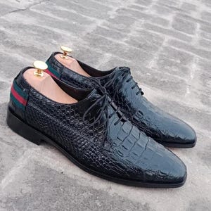 K&ouml;nnte beinhalten: Ein Paar schwarze Lederschuhe mit einem strukturierten, krokodilartigen Muster. Die Schuhe haben Schn&uuml;rsenkel und ein gr&uuml;n-rotes Streifendetail an der Ferse. Schuhspanner sind eingesetzt.