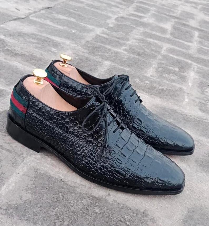 K&ouml;nnte beinhalten: Ein Paar schwarze Lederschuhe mit einem strukturierten, krokodilartigen Muster. Die Schuhe haben Schn&uuml;rsenkel und ein gr&uuml;n-rotes Streifendetail an der Ferse. Schuhspanner sind eingesetzt.