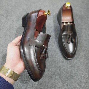 May include: A pair of dark brown leather loafers featuring a polished finish, a decorative strap across the vamp, and two small tassels. Each shoe includes a wooden shoe tree with a gold-toned metal knob inserted into the heel area.
