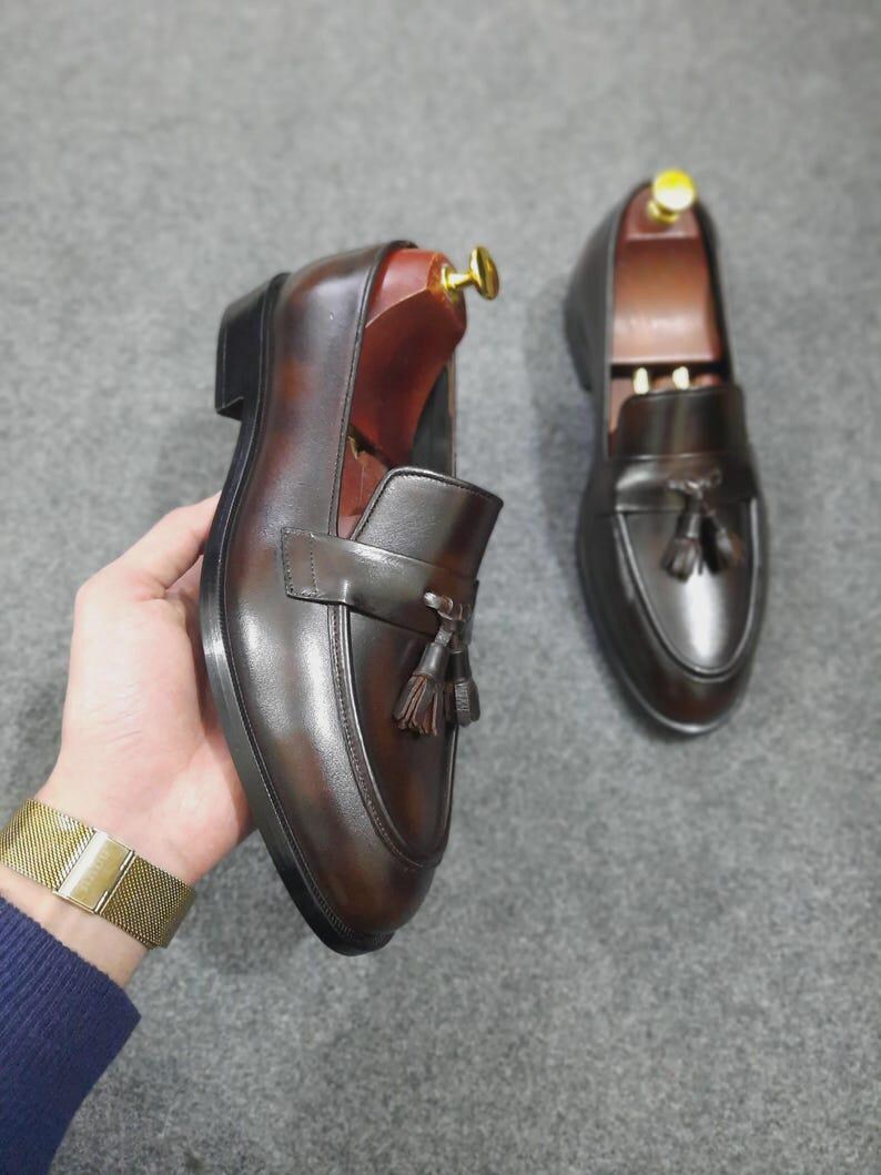 May include: A pair of dark brown leather loafers featuring a polished finish, a decorative strap across the vamp, and two small tassels. Each shoe includes a wooden shoe tree with a gold-toned metal knob inserted into the heel area.