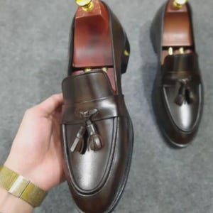 May include: A pair of dark brown leather loafers featuring a decorative tassel on the vamp and a smooth, polished finish. Each shoe is equipped with a wooden shoe tree insert that has a gold-colored metal knob at the heel for structural support.