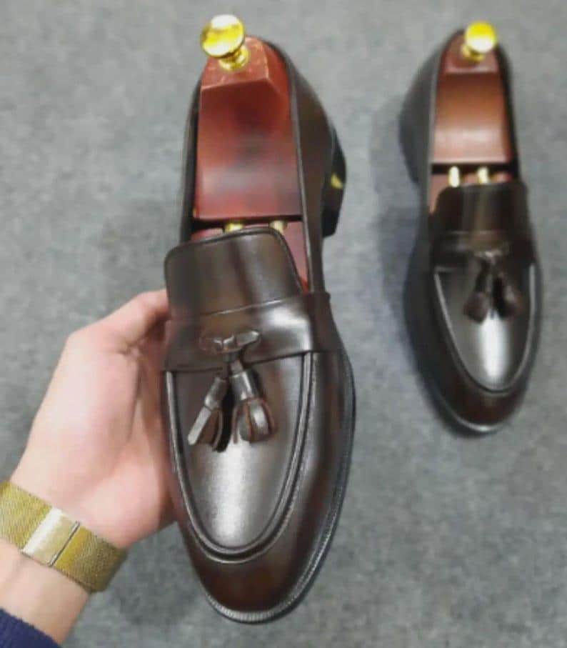 May include: A pair of dark brown leather loafers featuring a decorative tassel on the vamp and a smooth, polished finish. Each shoe is equipped with a wooden shoe tree insert that has a gold-colored metal knob at the heel for structural support.