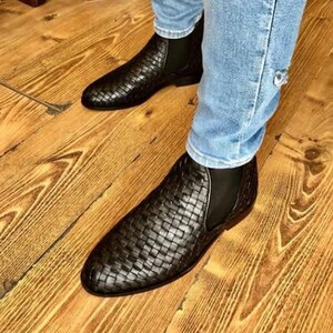 Op de afbeelding: Zwarte Chelsea-laarzen van geweven leer. De laarzen hebben een instapontwerp met elastische zijpanelen. De schoenen worden gedragen met een blauwe spijkerbroek. De laarzen staan op een houten vloer.