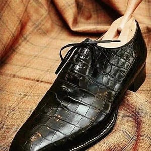 May include: A black leather dress shoe with a textured, crocodile-like pattern. The shoe has a pointed toe, a lace-up closure, and a wooden shoe tree inserted inside. The shoe is set on a brown plaid fabric.