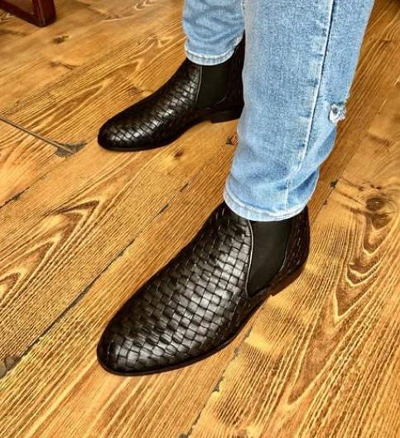 Op de afbeelding: Zwarte Chelsea-laarzen van geweven leer. De laarzen hebben een instapontwerp met elastische zijpanelen. De schoenen worden gedragen met een blauwe spijkerbroek. De laarzen staan op een houten vloer.