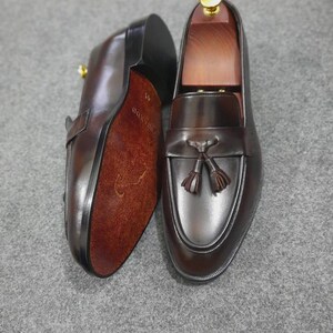May include: A pair of dark brown leather tassel loafers with a smooth finish. One shoe is positioned to show the textured brown sole with the size 41 printed on it, while the other displays the top with a decorative tassel and a wooden shoe tree.