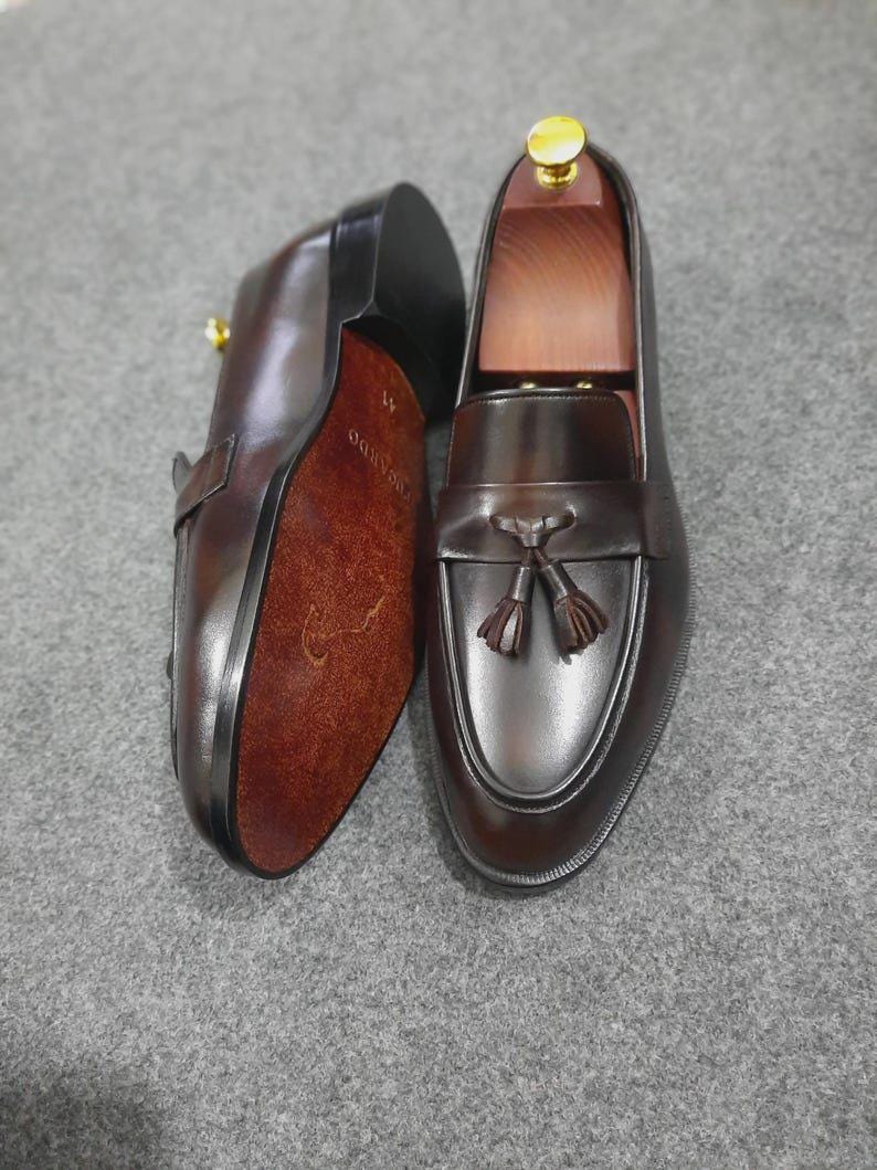 May include: A pair of dark brown leather tassel loafers with a smooth finish. One shoe is positioned to show the textured brown sole with the size 41 printed on it, while the other displays the top with a decorative tassel and a wooden shoe tree.