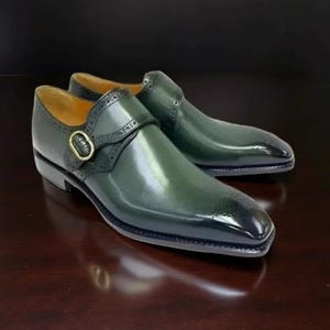 K&ouml;nnte beinhalten: Ein Paar gr&uuml;ne Lederschuhe mit spitzer Zehenpartie und Schnallenverschluss. Die Schuhe haben dekorative Perforationen und eine goldfarbene Schnalle. Die Schuhe werden auf einer dunkelbraunen Oberfl&auml;che pr&auml;sentiert.